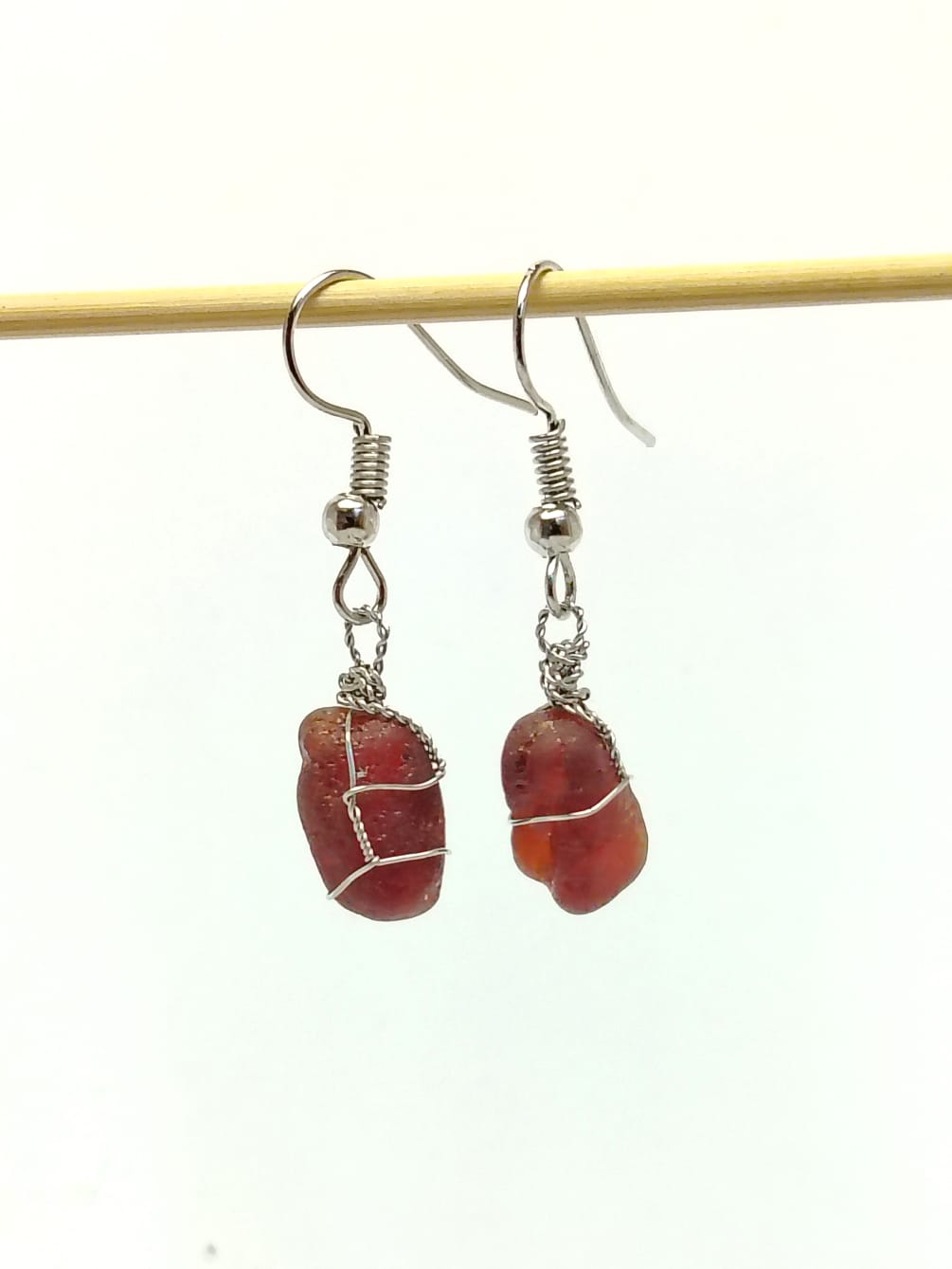A pair of handmade dangle earrings featuring rough, polished reddish-brown stone pendants wrapped in silver wire, suspended from silver fishhook wires, hanging from a thin wooden stick against a plain white background.