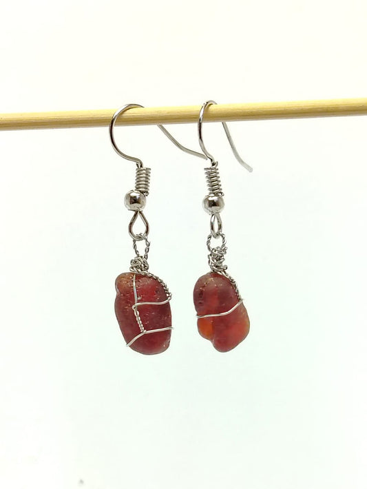 A pair of handmade dangle earrings featuring rough, polished reddish-brown stone pendants wrapped in silver wire, suspended from silver fishhook wires, hanging from a thin wooden stick against a plain white background.